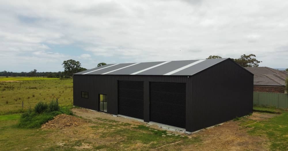 Black Australian-made steel shed in a grassy field with sliding windows, roller doors, and a skillion roof.