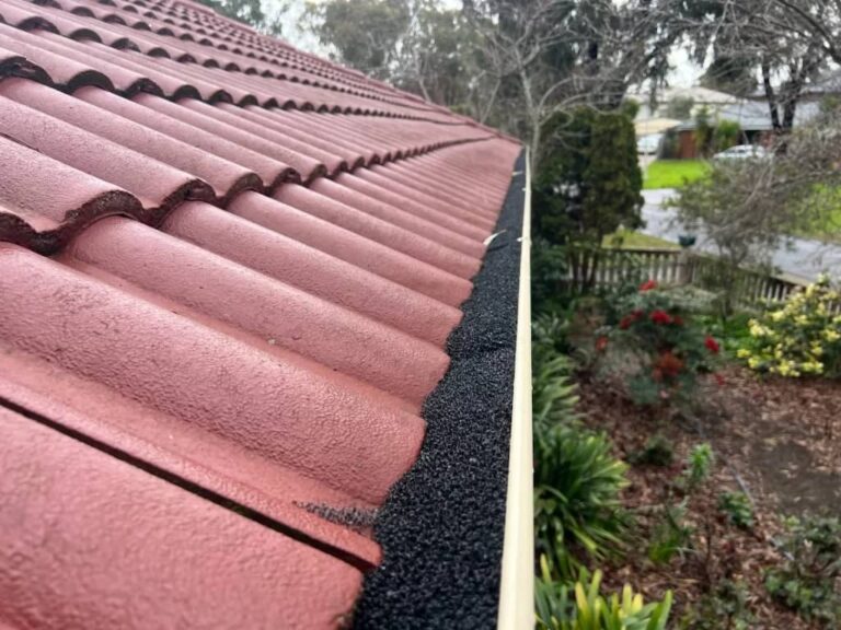 Gutter with installed brush guards on a red tile roof surrounded by a garden.