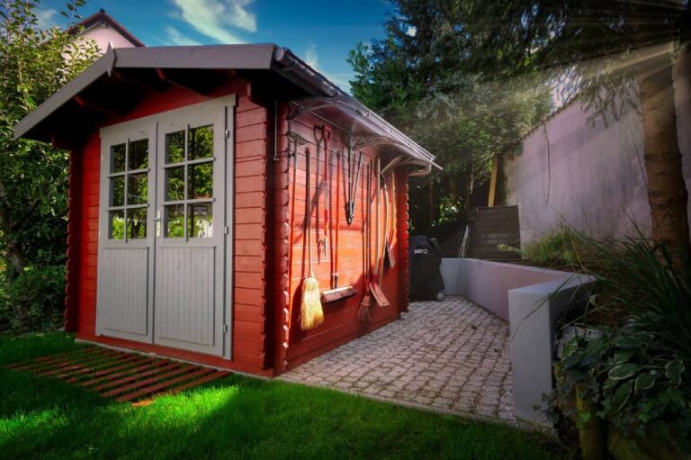 Red wooden storage shed with tools hanging on the side and a clean backyard, showcasing a neat and organized outdoor space.